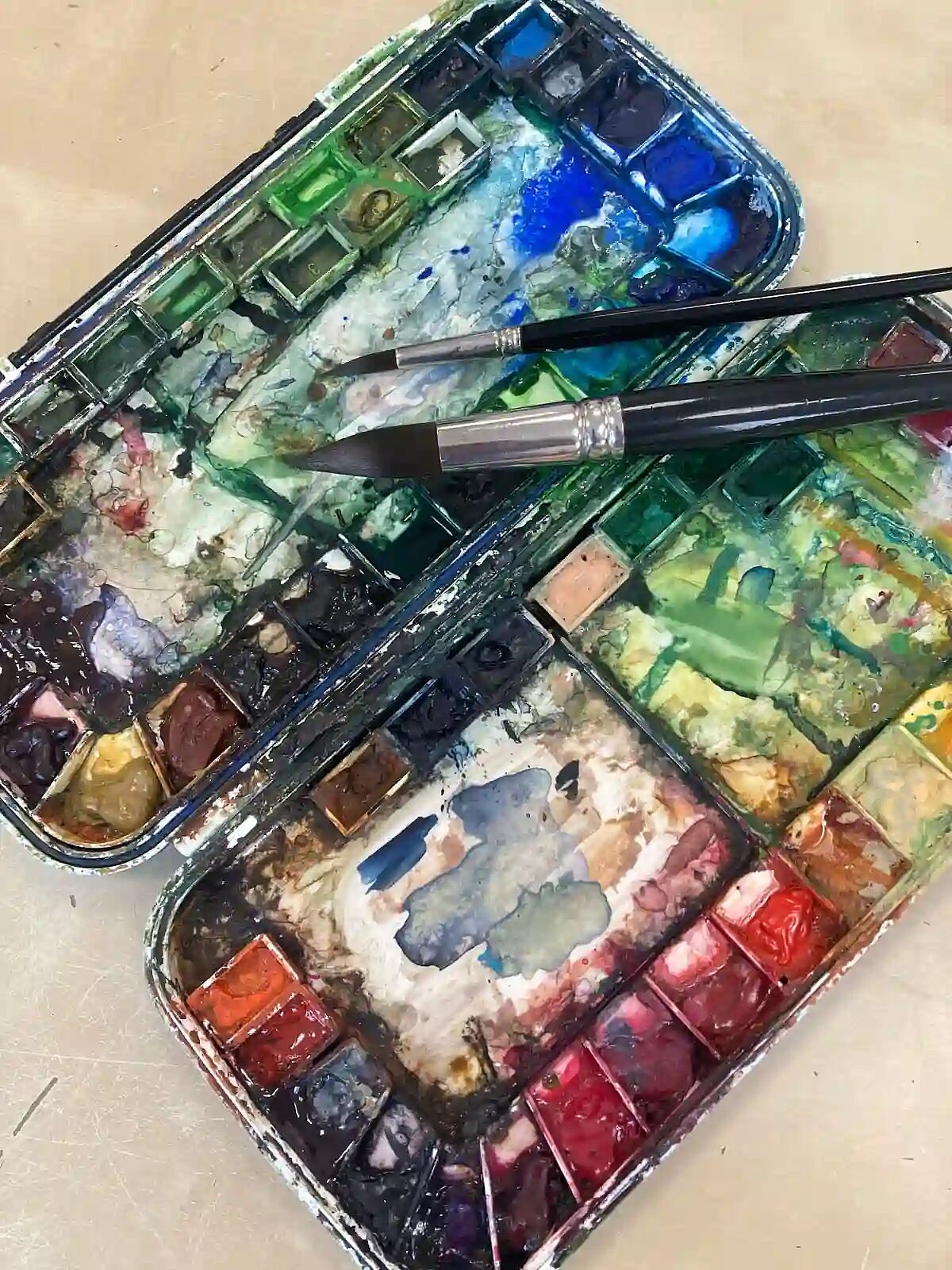 Das Bild zeigt einen geöffneten, gefüllten Aquarellmalkasten mit getrockneten Wasserfarben und Mischflächen sowie zwei feinen Pinseln.