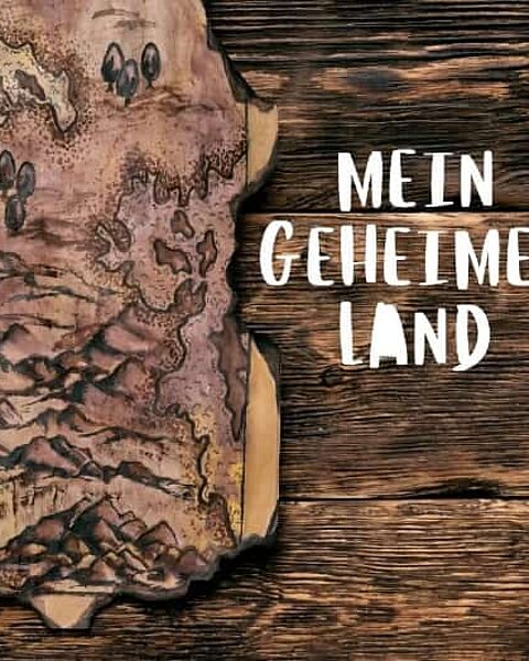 Die Grafik zeigt eine stilisierte, verbrannte Pergament- oder Schatzkarte auf einem dunklen Holzuntergrund mit dem deutschen Schriftzug Mein geheimes Land.